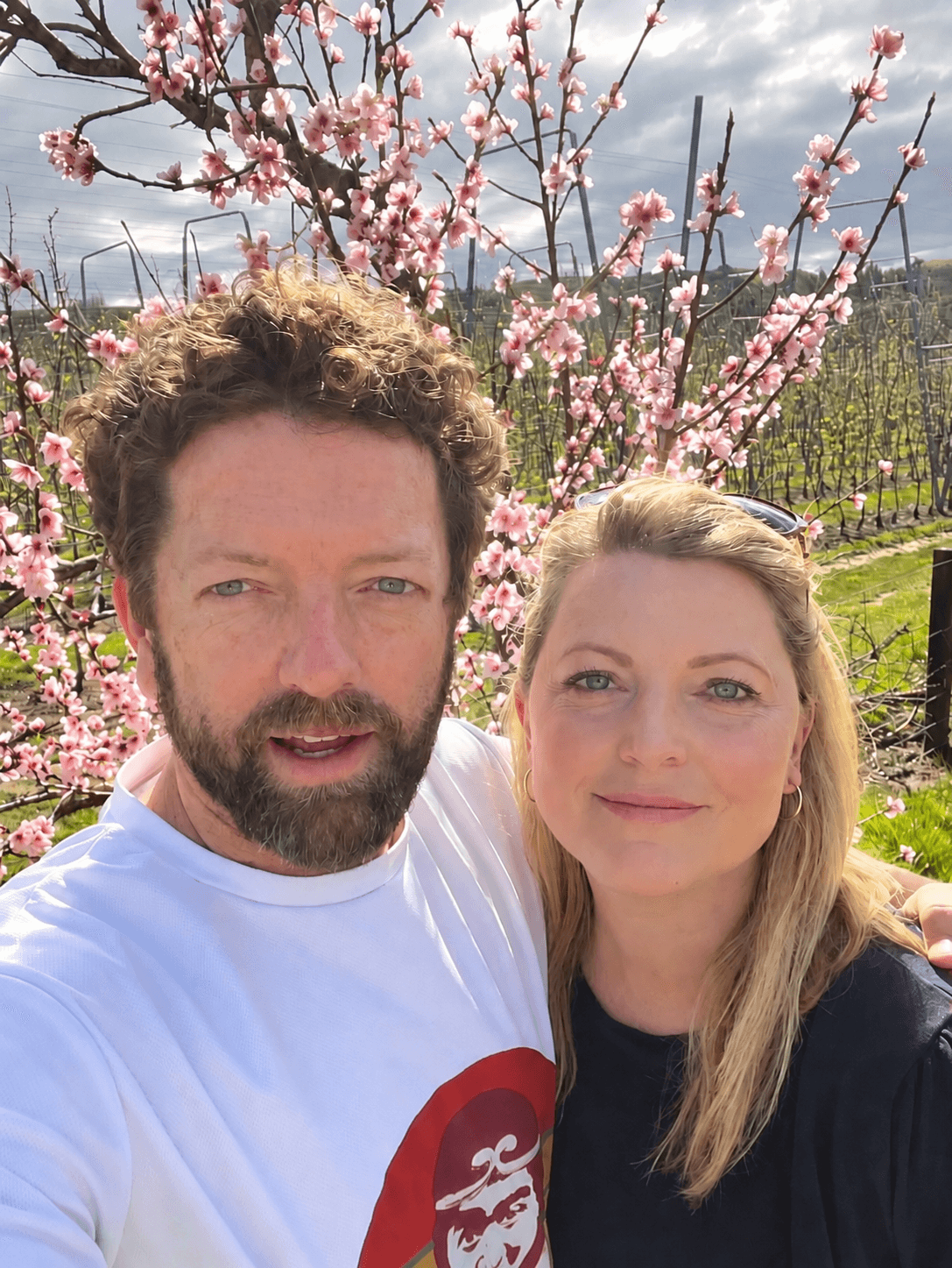 Bearded man and blonde woman smiling in front of blooming pink trees in an orchard.