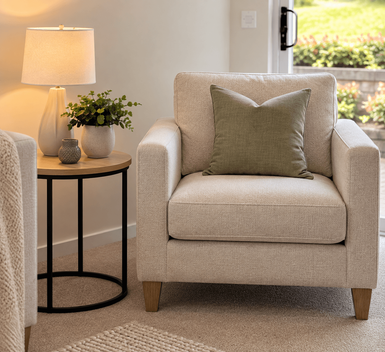 Beige armchair with an olive green pillow beside a wooden side table and lit lamp.