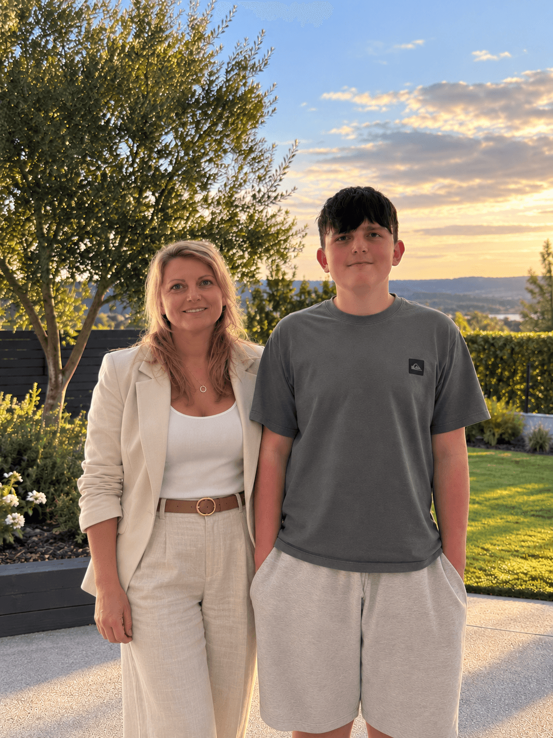 A woman and teenage boy pose outdoors against a scenic sunset with golden lighting.